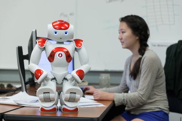 A NAO humanoid robot on a desk while a student works at a computer