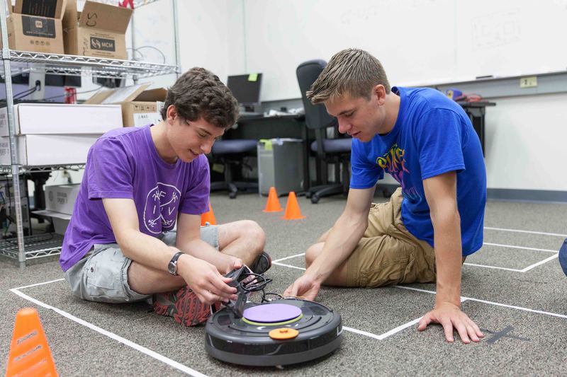 Two students on the lab floor setting up a small wheeled robot inside a taped-off course