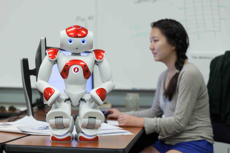 A NAO humanoid robot on a desk while a student works at a computer